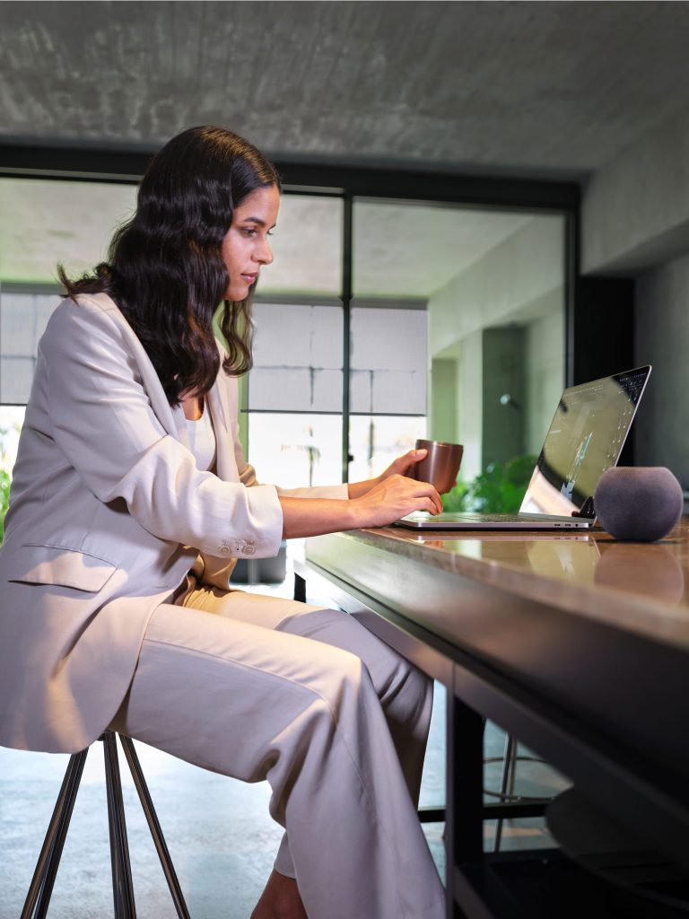 Woman sitting a home office desk on her laptop. The blinds on the window in the background are translucent and half way down.